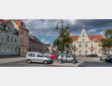 Ein gepflasterter Platz mit geparkten Autos, umgeben von historischen Gebäuden mit hellen Fassaden. Im Hintergrund steht ein gelbes, zweigeschossiges Rathaus mit Uhrturm und rotem Ziegeldach, daneben befinden sich bunte Wohnhäuser.