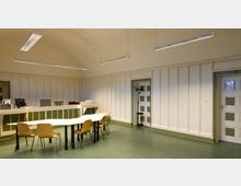 Ein einfacher Innenraum mit einer Empfangstheke und einem weißen Tisch mit mehreren Holzstühlen in der Mitte; die Wände sind beige mit vertikalen Vertiefungen, und es gibt grüne Akzente an Türen und Möbeln. Hängende LED-Leuchten beleuchten den Raum, und ein Garderobenständer steht in einer Ecke.