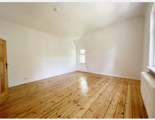 Heller, leerer Raum mit Holzdielenboden, weißen Wänden und einem Fenster mit Blick nach draußen; eine Holztür und ein Heizkörper sind ebenfalls sichtbar.