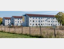 Mehrstöckiges Gebäude mit weißer Fassade und roten Ziegeldächern, umgeben von einem Zaun und einer Wiese im Vordergrund. Einige Bäume mit Herbstlaub stehen im Hintergrund.