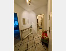 Flur in einer Wohnung mit heller Beleuchtung, einem weißen Teppich mit Rautenmuster und einem schmalen Konsolentisch an der Wand. Rechts eine gepolsterte Bank mit buntem Stoff und der Zugang zu einem Badezimmer, links ein Durchgang zu einem weiteren Raum mit blauem Licht.