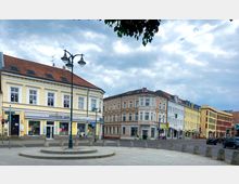 Stadtszene mit mehrstöckigen Gebäuden im klassischen Baustil und Geschäften im Erdgeschoss, darunter ein Optiker und ein Schuhgeschäft. Im Vordergrund ein gepflasterter Platz mit einer kleinen runden Plattform und einer Straßenlaterne, im Hintergrund eine belebte Straßenkreuzung unter bewölktem Himmel.