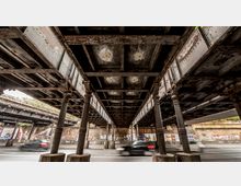 Unterseite einer alten Stahlbrücke mit dekorativen Metallsäulen, die über eine Straße führt. Im Hintergrund sind eine Backsteinmauer mit Graffiti sowie ein Gehweg sichtbar; Autos fahren auf der Straße darunter.