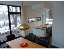 Moderne Wohnküche mit offenem Grundriss, einer Kücheninsel mit Spüle und Sitzecke mit Holztisch und schwarzen Stühlen. Große bodentiefe Fenster bieten Blick auf eine schneebedeckte Außenlandschaft mit Bäumen.