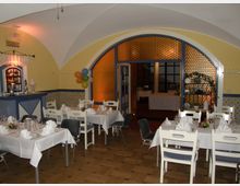 Ein gemütlicher Veranstaltungsraum mit gelb gestrichenen Wänden und Gewölbedecke, eingerichtet mit weiß eingedeckten Tischen und weißen Stühlen. Im Hintergrund befindet sich eine große Tür mit blauem Rahmen und Glasverkleidung, die zu einem weiteren Raum führt, dekoriert mit Ballons und stilisierten Wandornamenten.