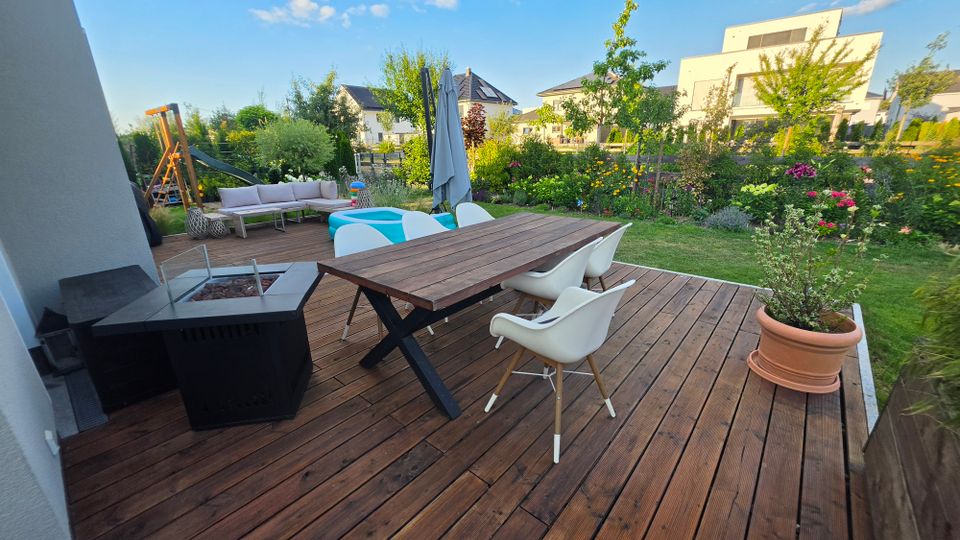 Holzterrasse in einem Wohnhausgarten mit großem Esstisch und weißen Stühlen, Feuerstelle, Loungesofa sowie Sonnenschirm und kleinem Kinderpool. Dahinter liegen Rasen, Blumenbeete und Bäume; im Hintergrund sind moderne Einfamilienhäuser zu sehen.