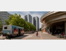 Straßenszene mit modernen Hochhäusern im Hintergrund, links eine Baumreihe, rechts ein Gebäude mit abgerundeten Metallverkleidungen und Geschäften. Im Vordergrund stehen ein bunt bemalter Transporter, Fußgänger und einige Fahrräder.