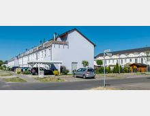 Reihenhaussiedlung mit weiß verputzten Gebäuden, mehreren Carports und parkenden Autos. Straßenschilder mit den Namen "Lebensstraße" und "Nebelstraße" stehen an einer Straßenecke, umgeben von Gepflegten Vorgärten und teilweise sichtbaren Wohngebäuden im Hintergrund.