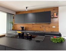 Moderne Küche mit dunklen Schränken und Arbeitsflächen in Schwarz sowie einer Rückwand aus warmem Holz. Im Vordergrund befindet sich eine Kücheninsel mit Kochfeldern, Kochgeschirr und frischen Zutaten.