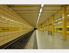 Untergrundbahnhof mit gelb gefliesten Wänden und gelben Säulen, der mit der Aufschrift „Weberwiese“ gekennzeichnet ist. Ein gelber Zug ist teils sichtbar, während er den Bahnhof passiert, neben einem Bahnsteig mit Sitzbänken und ausgeschilderten Informationen.