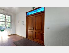 Eingangsbereich mit einer großen, doppelflügeligen Holztür mit Oberlicht aus blauen Glaselementen. Links daneben befindet sich ein Fenster mit weißen Rahmen, ein Heizkörper und eine grüne Gießkanne, der Boden ist mit roten Fliesen ausgelegt.