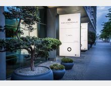 Moderner Hoteleingang mit großen beleuchteten Werbeschildern mit der Aufschrift „The Mandala Hotel“. Im Vordergrund stehen dekorative, in großen Pflanzgefäßen begrünte Bäume und Pflanzen entlang eines gepflasterten Gehwegs.