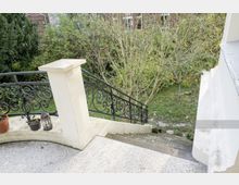 Eine Außentreppe mit hellem Steinbelag und einem dekorativen schwarzen Eisen-Geländer, die zu einem Garten mit Bäumen und Rasenfläche führt. Im Vordergrund befinden sich Blumenkübel und eine Laterne auf der Brüstung.