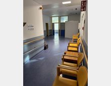 Flur in einem Krankenhaus oder einer medizinischen Einrichtung mit blauem Boden, weißen Wänden und Geländern an den Seiten. Rechts stehen gelbe Stühle mit Holztischen in einer Wartebereichs-Anordnung, während am Ende des Flurs Türen und Fenster zu sehen sind.