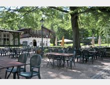 Ein Biergarten mit aufgestellten Tischen und Stühlen auf einem Kiesboden, umgeben von Bäumen. Im Hintergrund ein Gebäude im traditionellen Stil mit Holzfassade und Blumenkästen an den Fenstern.