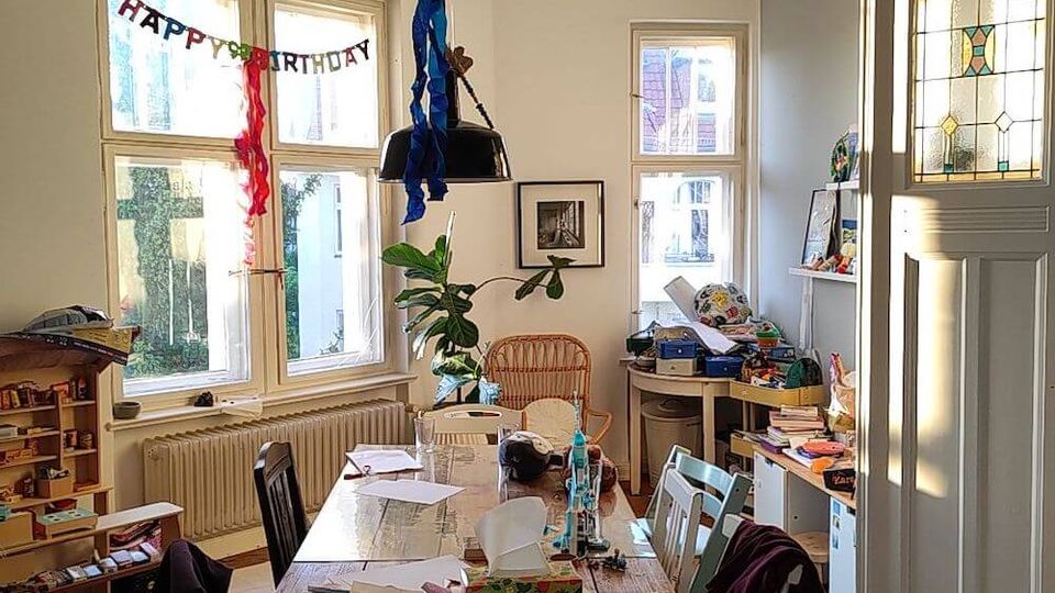 Dining room with a large wooden table covered in papers and household items, surrounded by mismatched chairs. The space features a ceiling light with colorful streamers, stained glass on a door, and "Happy Birthday" decorations on the windows.
