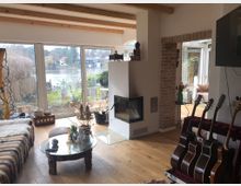 Wohnzimmer mit Blick auf einen Garten und einen See durch große Fenster. Der Raum enthält eine Sitzecke mit einem Couchtisch aus Glas, einen modernen Kamin, eine offene Backsteinwand und mehrere Gitarren in einem Ständer.