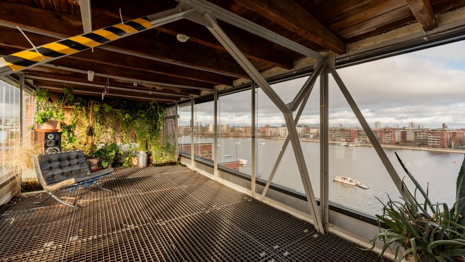 Industrieartig gestalteter Innenraum mit Gitterrostboden, sichtbaren Stahlträgern und Holzbalkendecke sowie einer begrünten Pflanzenwand. Große Fensterfront mit Blick auf einen breiten Fluss und gegenüberliegende Stadtbebauung.