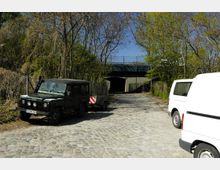 Ein gepflasterter Weg führt unter eine Brücke, flankiert von Bäumen und einem Maschendrahtzaun. Links steht ein dunkler Geländewagen mit Anhänger, rechts ist ein weißer Transporter zu sehen.