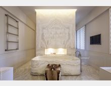 Modernes Badezimmer mit einer freistehenden Steinbadewanne vor einer Wand aus beigem Marmor mit symmetrischer Maserung. An der Wand befindet sich eine beleuchtete Nische, rechts ein kleiner Fernseher, und links ein verchromter Handtuchhalter.