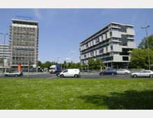 Offene Grünfläche mit Blick auf eine belebte Kreuzung, gesäumt von zwei modernen Bürogebäuden. Das linke Gebäude trägt ein „HypoVereinsbank“-Logo und ist teilweise von einem Baugerüst bedeckt, während das rechte Gebäude eine mehrdimensionale Fassade mit großen Fenstern aufweist; im Hintergrund sind Bäume sichtbar.