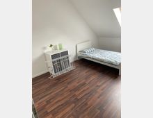 Schlicht eingerichtetes Schlafzimmer mit einem Bett mit weißem Rahmen und gestreifter Bettwäsche, einer weißen Kommode mit dunklen Schubladen, einer Blumenvase und einem Trockengestell. Der Raum hat einen dunklen Holzfußboden und eine Dachschräge mit einem großen Dachfenster.