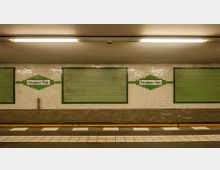 U-Bahn-Station mit einer Wand aus weißen Fliesen, auf der "Potsdamer Platz" in einem grünen Fliesenrahmen steht. Zwei grüne, rechteckige Flächen flankieren die Beschilderung, der Bahnsteigbereich ist mit weißen und grauen Bodenfliesen gestaltet.