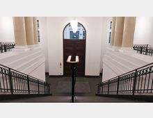 Ansicht eines symmetrischen Treppenhauses mit schwarzen Metallgeländern, weißen Wänden und großen beigen Säulen. Im Hintergrund befindet sich eine doppelflügelige dunkle Holztür mit einem halbkreisförmigen Fenster darüber.