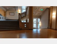 Treppenhaus in einem historischen Gebäude mit Steinbalustrade und verzierten Buntglasfenstern. Die Decke ist mit Kassettenmuster gestaltet, und die Wände sind in warmen Tönen gehalten, beleuchtet von Wandleuchten.