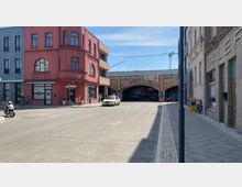 Straßenszene mit einem roten Eckgebäude, auf dem „Hotel“ steht, flankiert von weiteren Gebäuden. Im Hintergrund eine Bahnunterführung aus Backstein mit Graffiti, auf der Baustellenkräne sichtbar sind, sowie geparkte Fahrräder und ein Auto auf der Straße.