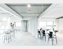 Heller, moderner Mehrzweckraum mit offenen Fensterfronten, mit hohen, weißen Tischen und Barhockern auf der linken Seite sowie schwarzen Stühlen mit Schreibflächen auf der rechten Seite. Der Raum ist durch weiße Vorhänge teilweise abgetrennt und hat ein freiliegendes Betondecken-Design.
