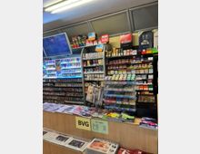 Ein kleiner Kiosk mit einem Verkaufstresen, darauf Zeitschriften, Zeitungen und Schilder zu SIM-Karten und Preisen. Im Hintergrund befinden sich Regale mit Tabakwaren, Filterprodukten und weiteren Waren, darunter Süßigkeiten und Zubehör.