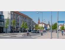 Straßenansicht einer urbanen Umgebung mit mehrstöckigen Wohn- und Geschäftsgebäuden, einem Gehweg mit Bäumen und Straßenlaternen sowie einer Kirche mit Turm im Hintergrund. Auf der Straße fahren Autos und Fahrräder, während Passanten den Gehweg nutzen; ein Straßenschild weist auf die "Goethestraße" hin.