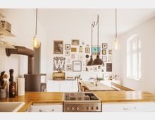 Helle Küche mit einer Kochinsel aus Holz, einem Elektroherd und einer Spüle im Vordergrund. Im Hintergrund befinden sich ein Esstisch, ein Kaminofen sowie eine Wand mit zahlreichen gerahmten Bildern und Dekorationen; seitlich ein Fenster mit Tageslicht.