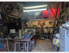 Eine Werkstatt mit unaufgeräumtem Innenraum, ausgestattet mit Werkbänken, Werkzeugen und verschiedenen Geräten. Die Wände sind alt und teilweise mit Neonlichtern und aufgehängten Fahrrädern dekoriert.