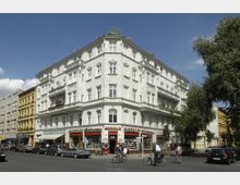 Eckgebäude mit historischer Fassade in Weiß und reich verzierten Fensterelementen, im Erdgeschoss befinden sich Geschäfte mit roten Markisen. Im Vordergrund sieht man eine Straße mit parkenden Autos, Radfahrern und Fußgängern, flankiert von weiteren Gebäuden und Bäumen.