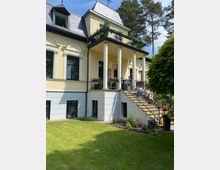 Gelbes, zweistöckiges Gebäude mit einer Veranda auf Säulen, darüber ein Balkon mit Blumen. Das Haus hat ein Dach mit Schieferdeckung, viele Fenster und ist von einem Garten mit Rasen und Bäumen umgeben.