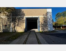 Ein großes Gebäude mit breitem, blauem Tor, das in eine dunkle Halle führt. Vor dem Eingang verlaufen Schienen und ein gepflasterter Weg, umgeben von Bäumen und Schatten.