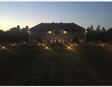 Ein beleuchtetes Herrenhaus bei Dämmerung, umgeben von Bäumen. Im Vordergrund befindet sich eine große Rasenfläche, die von kleinen Bodenlichtern gesäumt wird.