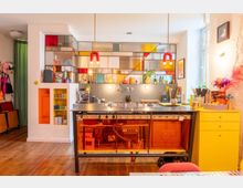 Bright, modern kitchen with a stainless-steel island and orange-tinted glass base, lit by two red pendant lights. A wall of open shelving with multicolored glass panels forms a backdrop above a gray tile backsplash, with a yellow chest of drawers and wooden floor in the foreground.