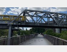 Eine Metallbrücke mit einer gelben U-Bahn darauf überspannt einen Fußgängerweg. Im Hintergrund sind Bäume, eine moderne Architektur und ein teilweise bewölkter Himmel zu sehen.