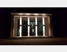 Modernes Gebäude mit rechteckigem Eingang, beleuchtet durch mehrere vertikale Lampen an Säulen. Die Glasfassade zeigt den Innenraum mit Treppen und Beleuchtung, während breite Treppen zum Eingang führen; die Szene ist nachts aufgenommen.