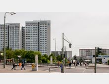 Wohnblock im Stadtgebiet mit mehreren Hochhäusern aus weißem Beton, umgeben von Bäumen und einer Hauptstraße im Vordergrund. Auf der Straße sind Fußgänger, Straßenlaternen, Verkehrsampeln und Bushaltestellen sichtbar.