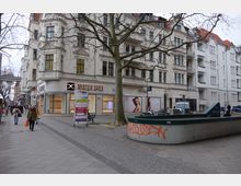Straßenszene mit einem mehrstöckigen Eckgebäude im Altbaustil, das Geschäfte wie „Mister Spex“ und „AktivOptik“ im Erdgeschoss beherbergt. Im Vordergrund sind ein Baum, ein mit Graffiti besprühter Betonrand sowie Fußgänger auf einem breiten Gehweg zu sehen.
