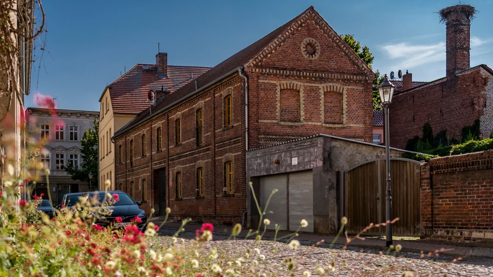 Blick auf eine ruhige Kopfsteinpflasterstraße mit einem zweigeschossigen Backsteingebäude mit Satteldach und zugemauerten Fenstern. Daneben stehen eine Garage mit Rolltor, eine Backsteinmauer und eine Straßenlaterne, im Vordergrund sind unscharfe Wildblumen zu sehen.