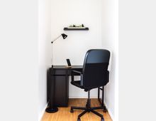 Schlichter Arbeitsbereich in einem kleinen, weißen Raum mit einem schwarzen Schreibtisch, einem schwarzen Bürostuhl auf Rollen, einer Stehlampe und einem Regal mit kleinen Pflanzen an der Wand. Der Boden besteht aus hellem Holz.