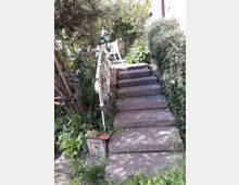 Steintreppe in einem Gartenbereich, umgeben von dichtem Grün, darunter Sträucher und Blumen. Ein weißes Metallgeländer verläuft neben der Treppe, und oben steht ein weißer Stuhl in einer sonnigen Ecke.