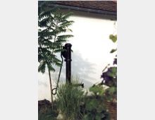 Ein alter, schwarzer Wasserpumpenbrunnen steht inmitten von hohen Gräsern und Pflanzen neben einer weiß verputzten Wand. Im Hintergrund ist ein Teil eines Ziegeldachs sichtbar, und ein Baum wirft Schatten auf die Wand.