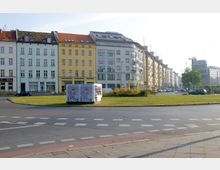 Straßenkreuzung mit Grünfläche in der Mitte, umgeben von Wohn- und Geschäftshäusern im klassischen und modernen Stil. Im Vordergrund ein großer Verteilerkasten mit Kunst darauf, sowie ein U-Bahn-Schild, das auf eine nahegelegene U-Bahn-Station hinweist.