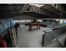 Industrieähnlicher Innenraum mit einer offenen Galerieebene, die mit Metallgeländern umgeben ist. Auf der Ebene befinden sich ein Tischkicker, schwarze Sitzwürfel, eine Wasserstation und Pflanzen, während die Konstruktion des Stahlträgers an der Decke sichtbar ist.